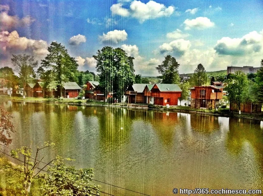 A lake surrounded by trees and wooden buildings, under a cloudy sky.