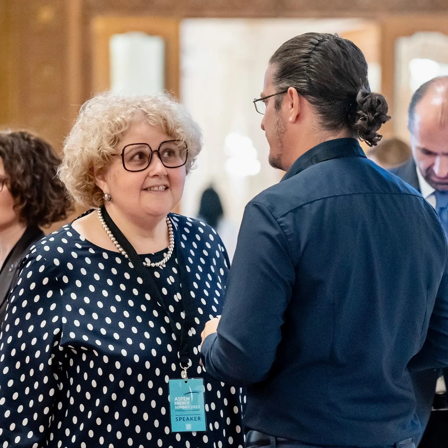 O femeie cu păr blond și creț, cu ochelari și o bluză cu buline, purtând o legitimație de vorbitor la Aspen Energy Summit 2023, discută cu un bărbat îmbrăcat într-o cămașă bleumarin.