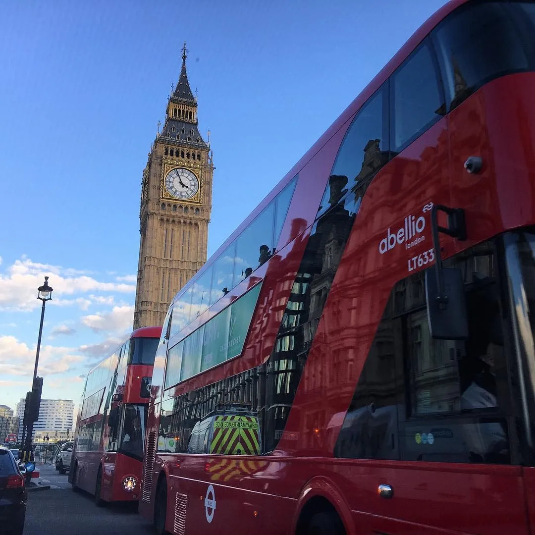 Autobuze roșii cu etaj pe o stradă din Londra, cu Big Ben înălțânduse în spatele lor pe fundalul unui cer albastru.