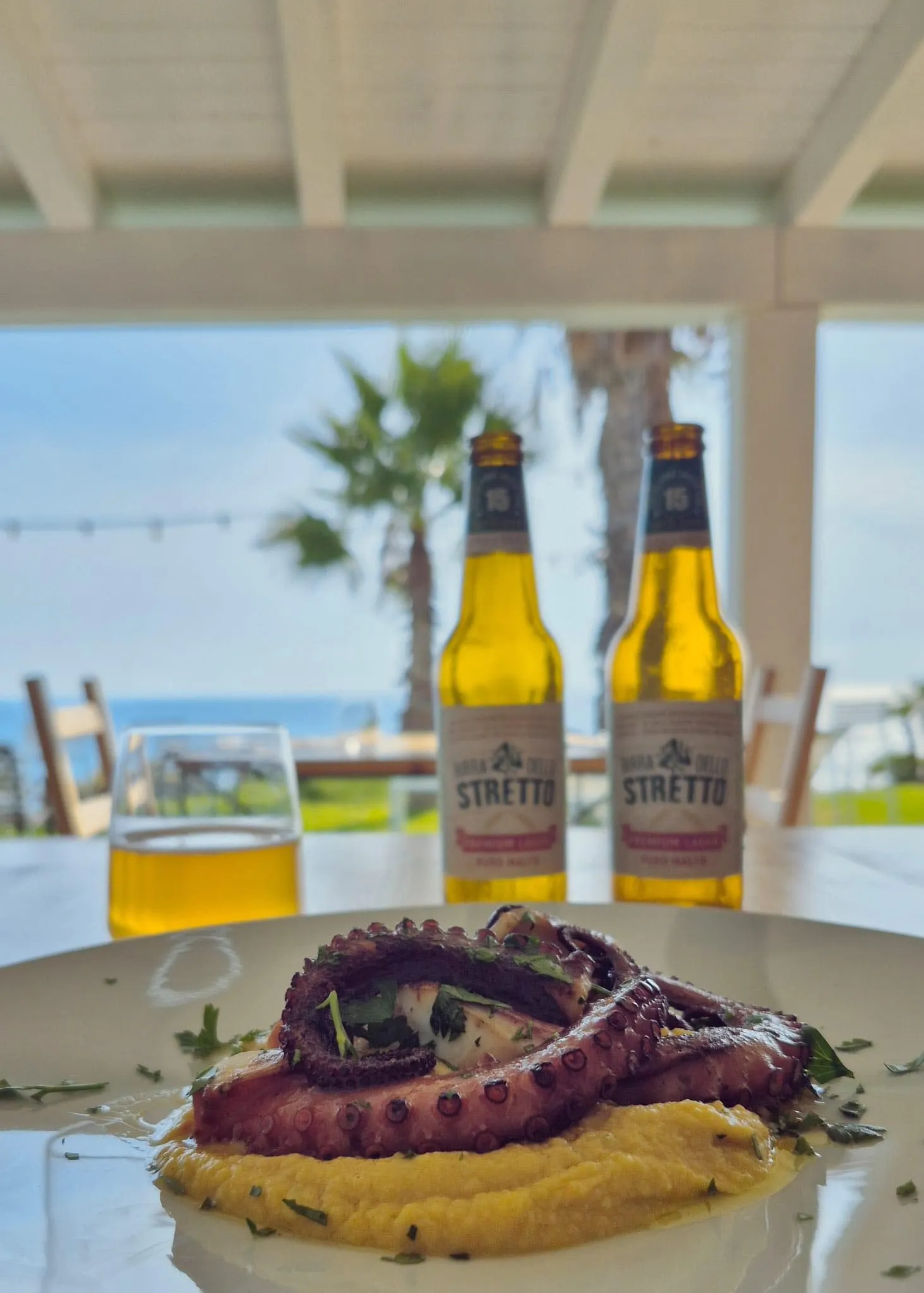 O farfurie cu caracatiță la grătar și două sticle de bere, pe masa unui restaurant de la malul mării.