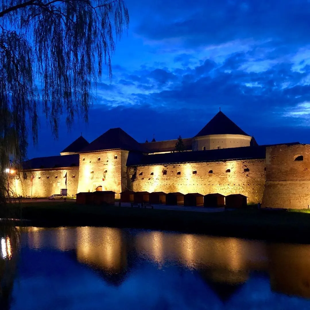 Illuminated medieval stone fortress with conical towers reflected in water under a deep blue dusk sky