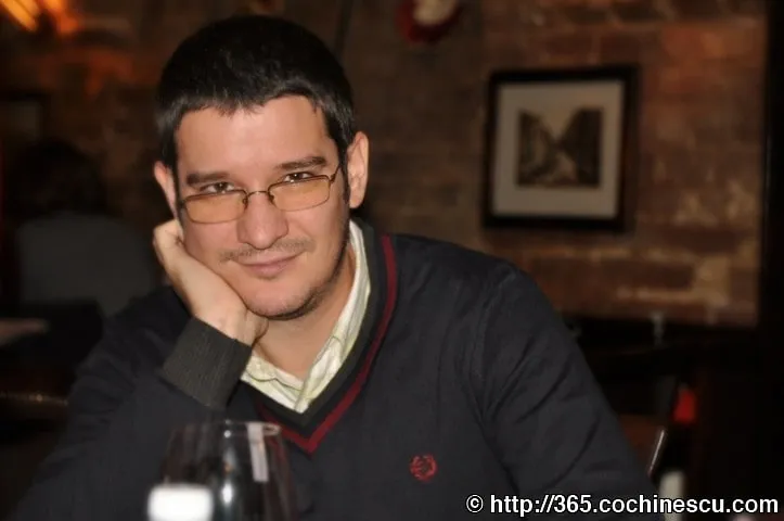 Un barbat cu ochelari zambeste sprijinindu-si capul in mana, la o masa.