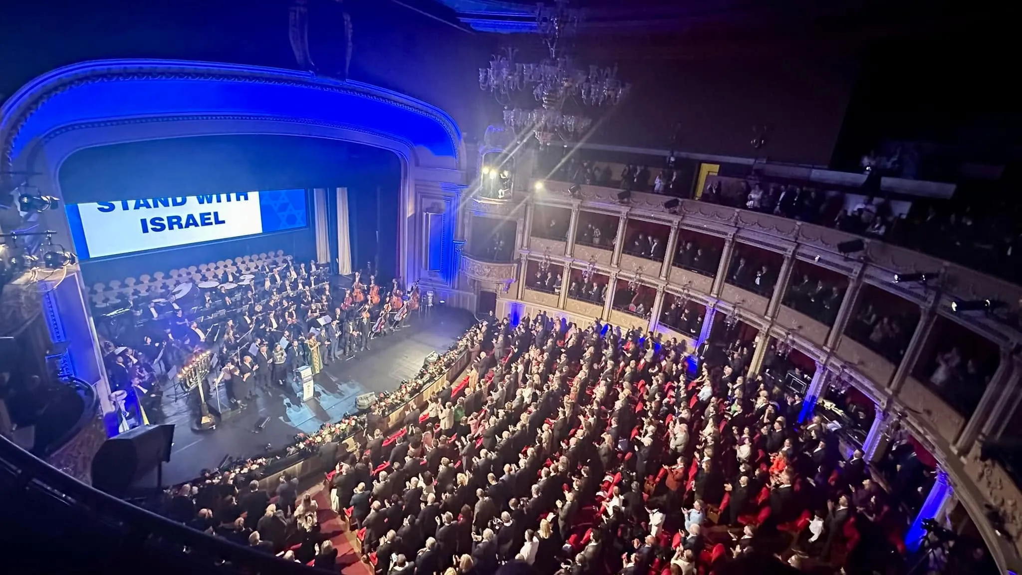 A packed ornate theater hall with an orchestra on stage beneath a large STAND WITH ISRAEL screen and balconies full of guests.