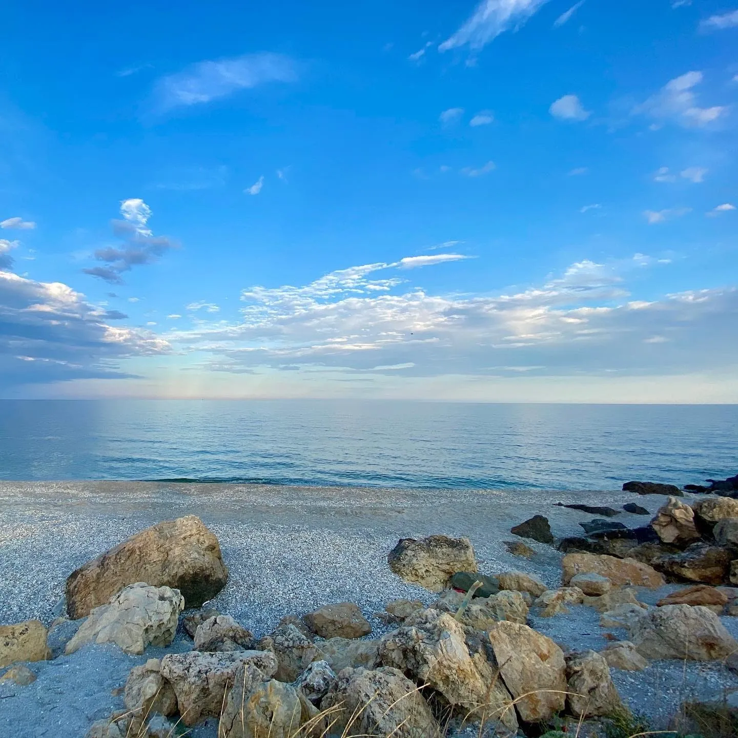 O plajă liniștită și pietroasă se întâlnește cu o mare albastră și calmă, sub un cer larg cu nori răzleți.