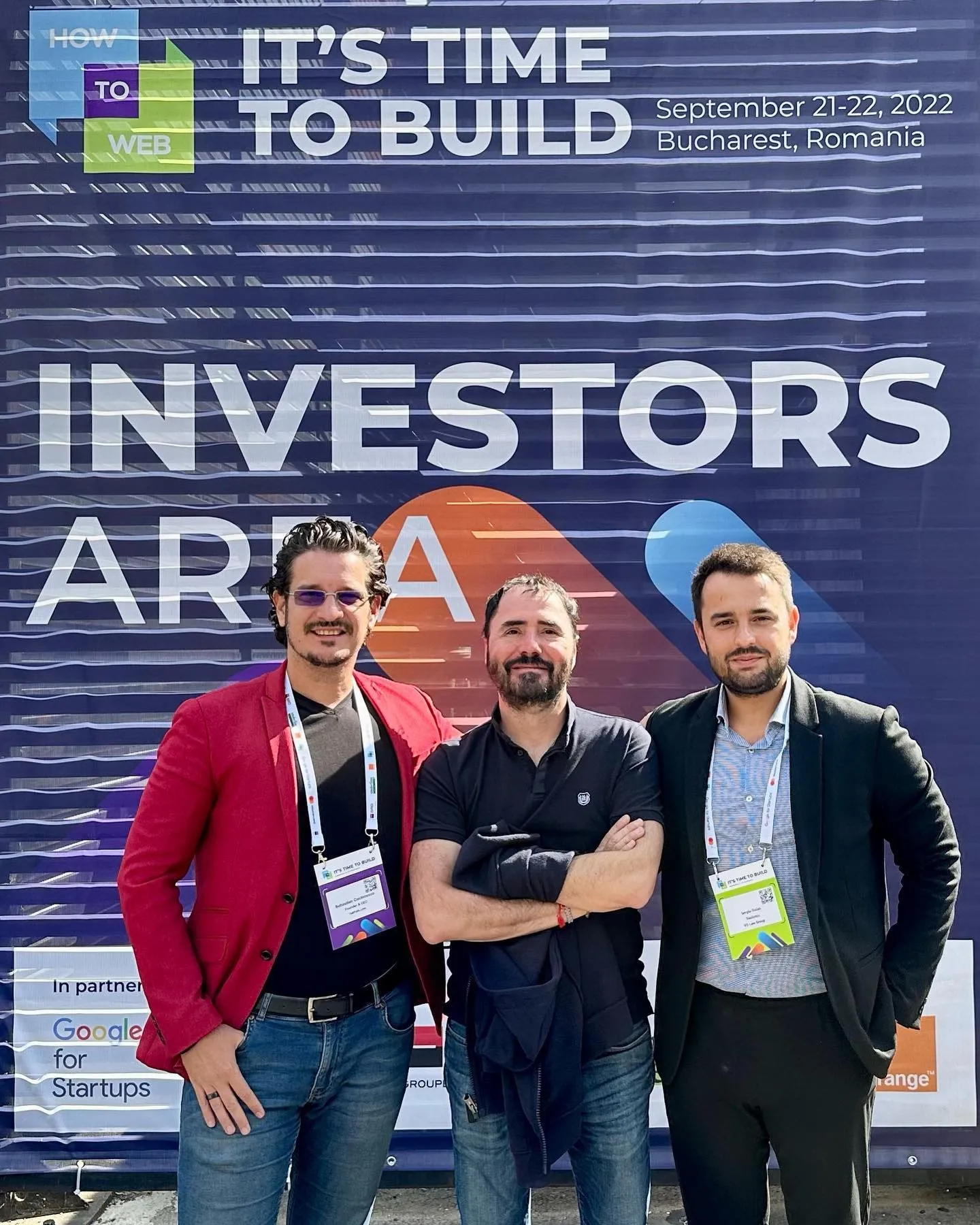 Three men pose together in front of the How to Web Investors Area banner in Bucharest.