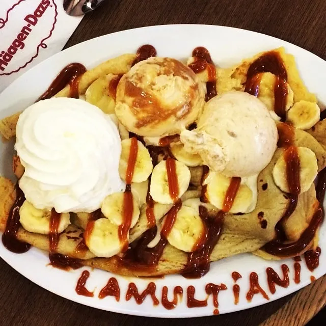 O farfurie cu clătite, banane, înghețată și frișcă, cu textul LA MULTI ANI scris cu sos pe margine.