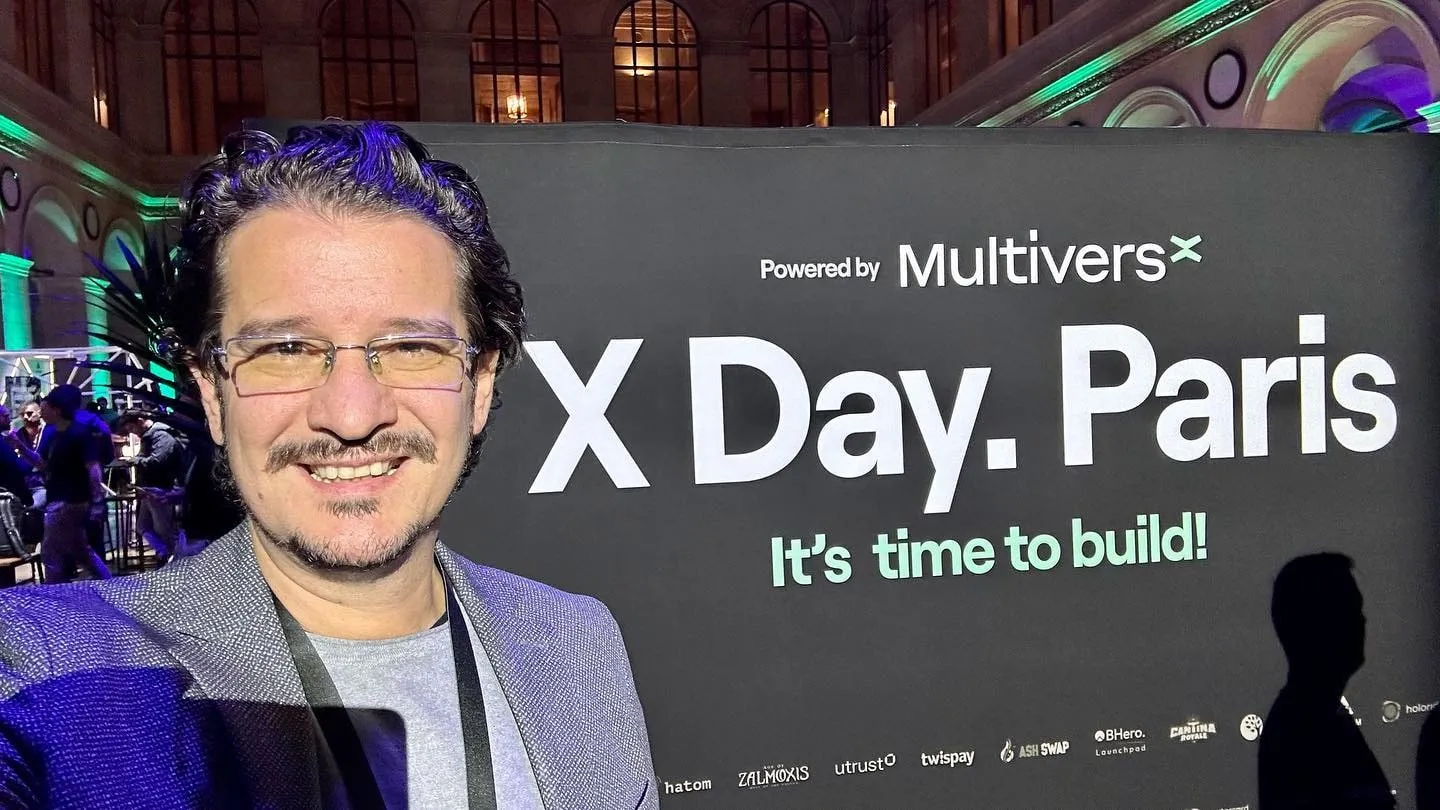 Bărbat cu ochelari zâmbind într-un selfie lângă un banner X Day Paris susținut de MultiversX.