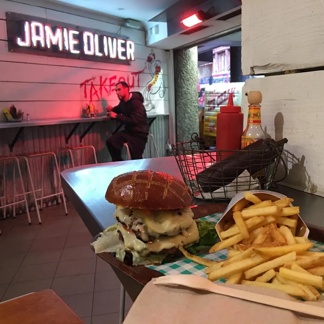 A cheeseburger and french fries on a table inside a Jamie Oliver takeout restaurant.