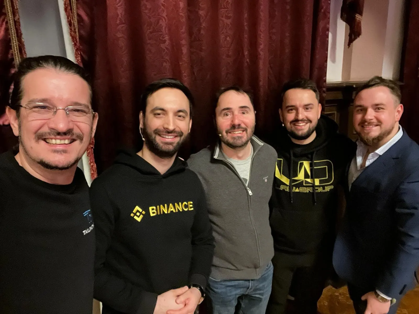 Cinci bărbați zâmbesc la o fotografie de grup făcută în interior, doi dintre ei purtând hanorace Binance și NU Group, în fața unor draperii roșii.