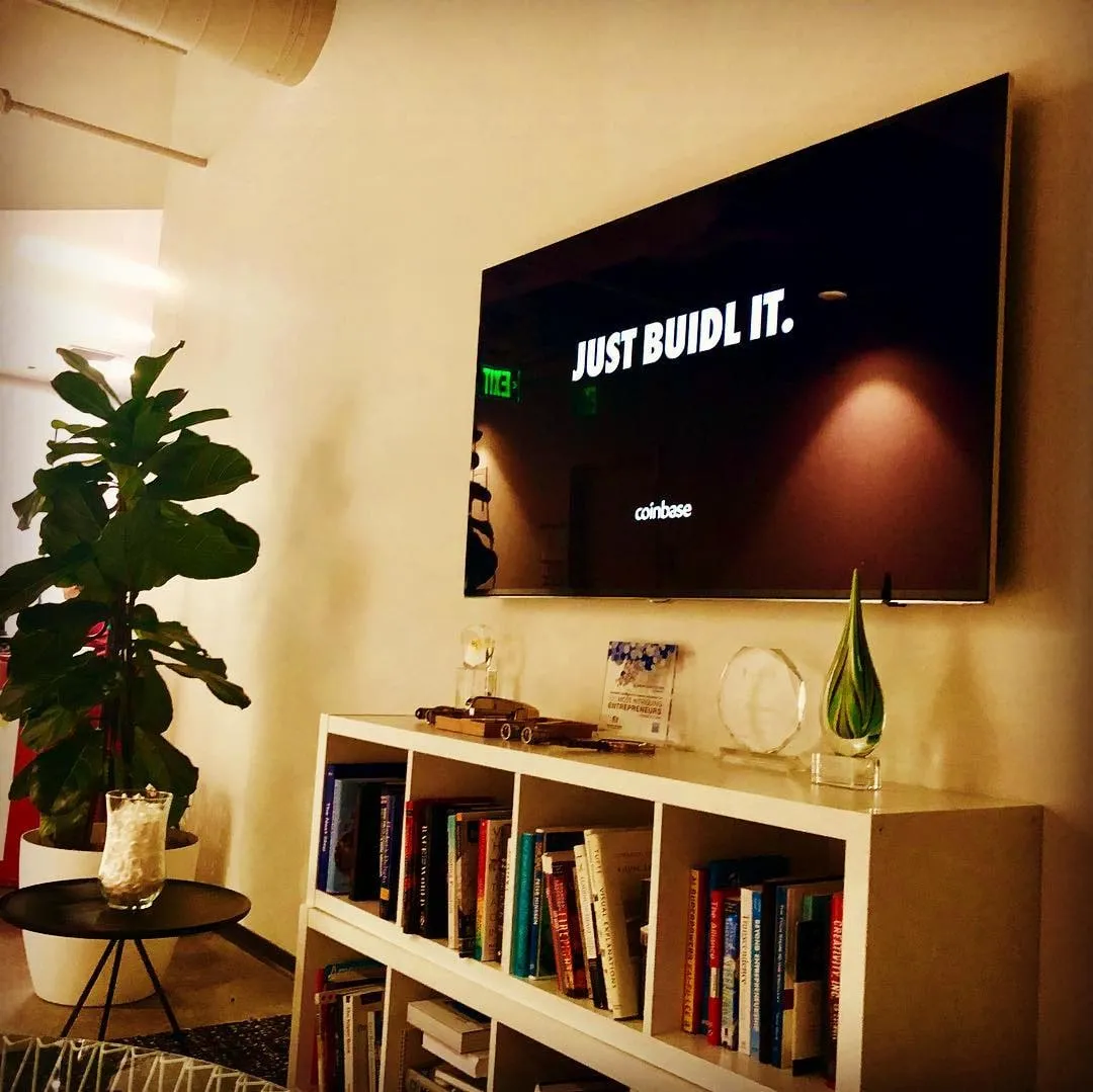 Televizor montat pe perete, pe care scrie Just BUIDL IT, și sigla Coinbase deasupra unei biblioteci albe cu premii și plante