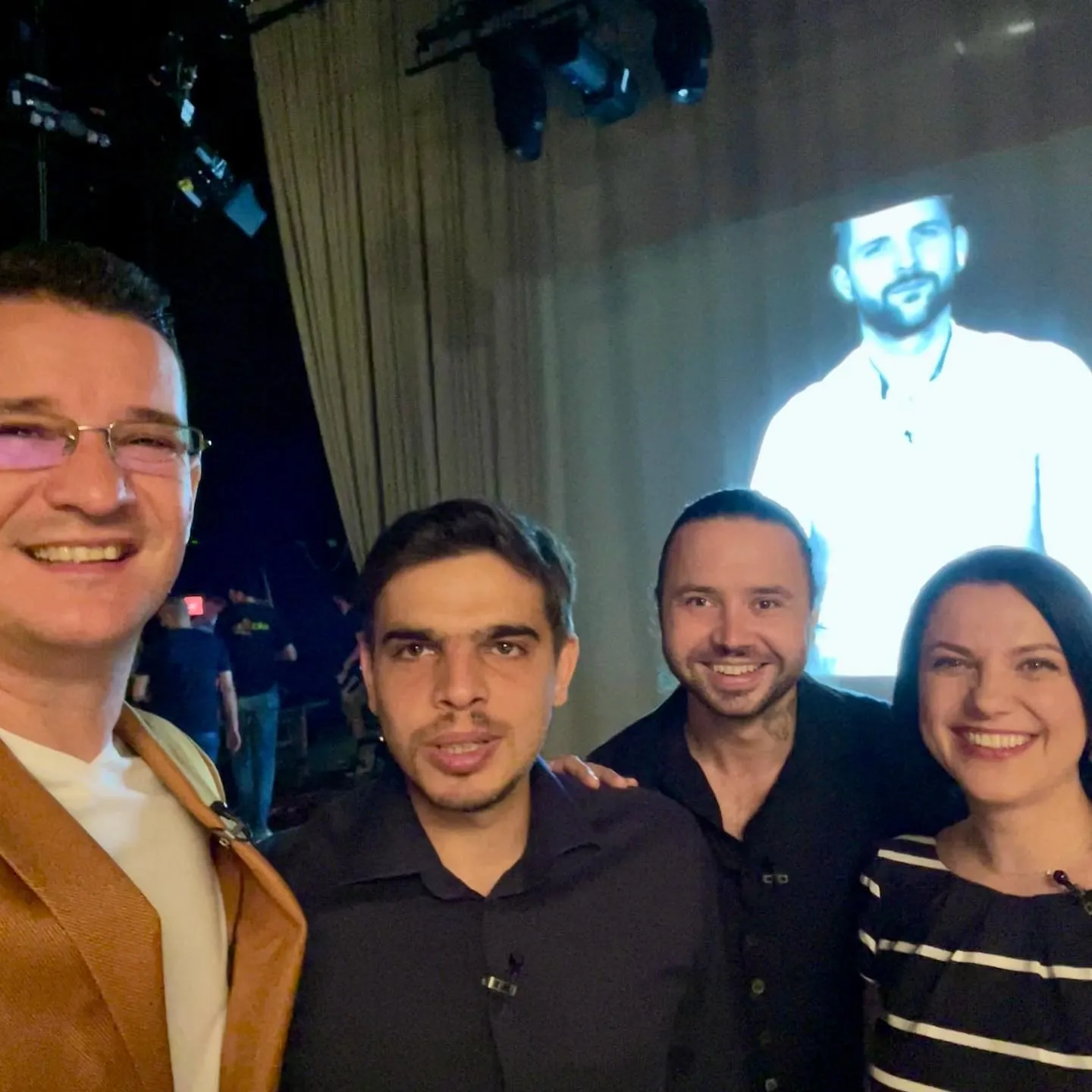 Patru persoane pozează pentru un selfie în culise, în fața unui ecran mare, luminat.