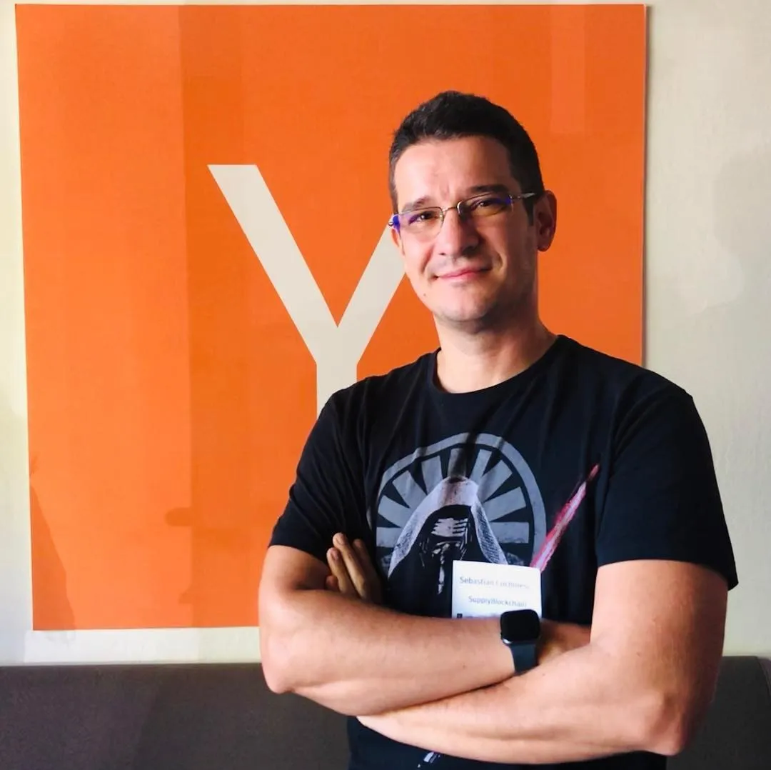 Bărbat într-un tricou negru cu imprimeu și ecuson, stând cu brațele încrucișate în fața unui logo Y Combinator mare și portocaliu
