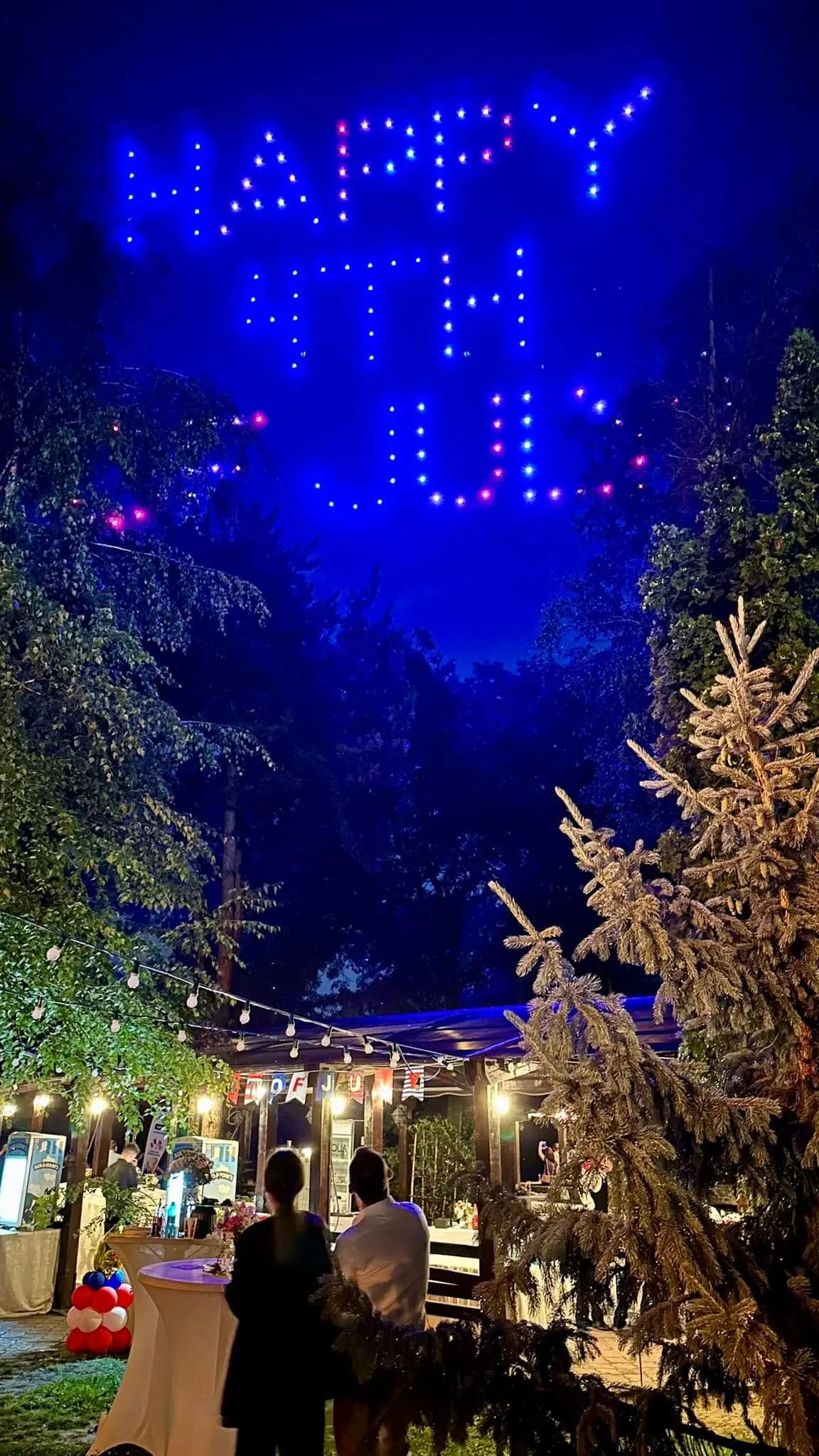 Night outdoor celebration with a drone light show spelling Happy 4th and guests under string lights with red, white, and blue balloons.