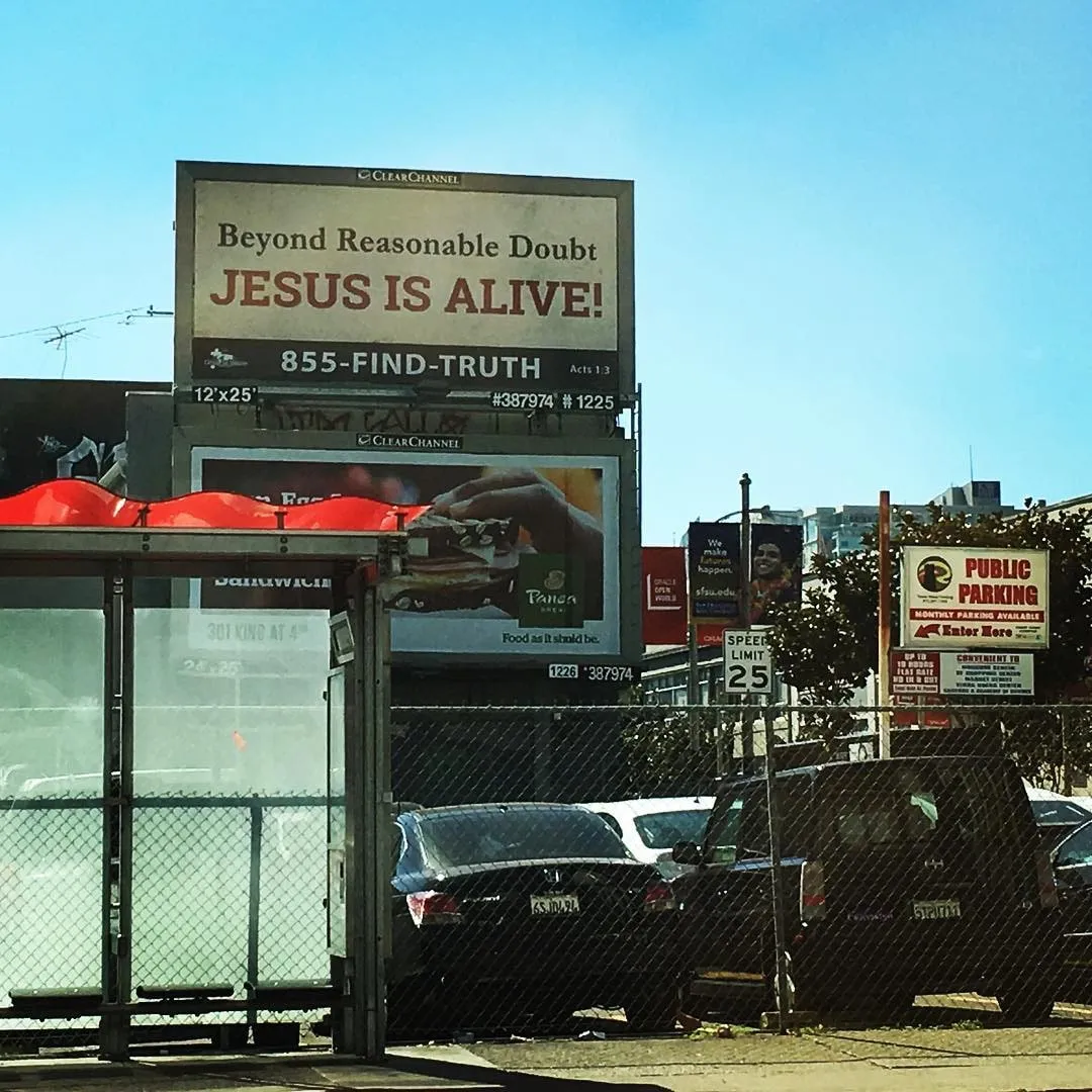 Billboard reading Beyond Reasonable Doubt JESUS IS ALIVE above a parking lot and bus shelter on a sunny day.