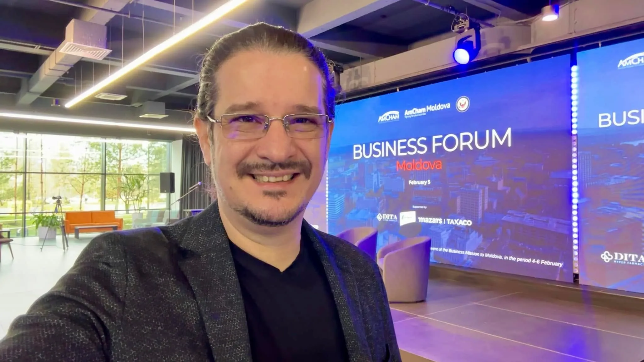 Sebastian Cochinescu zâmbește într-un selfie făcut la Business Forum Moldova, cu ecranul scenei în spate.