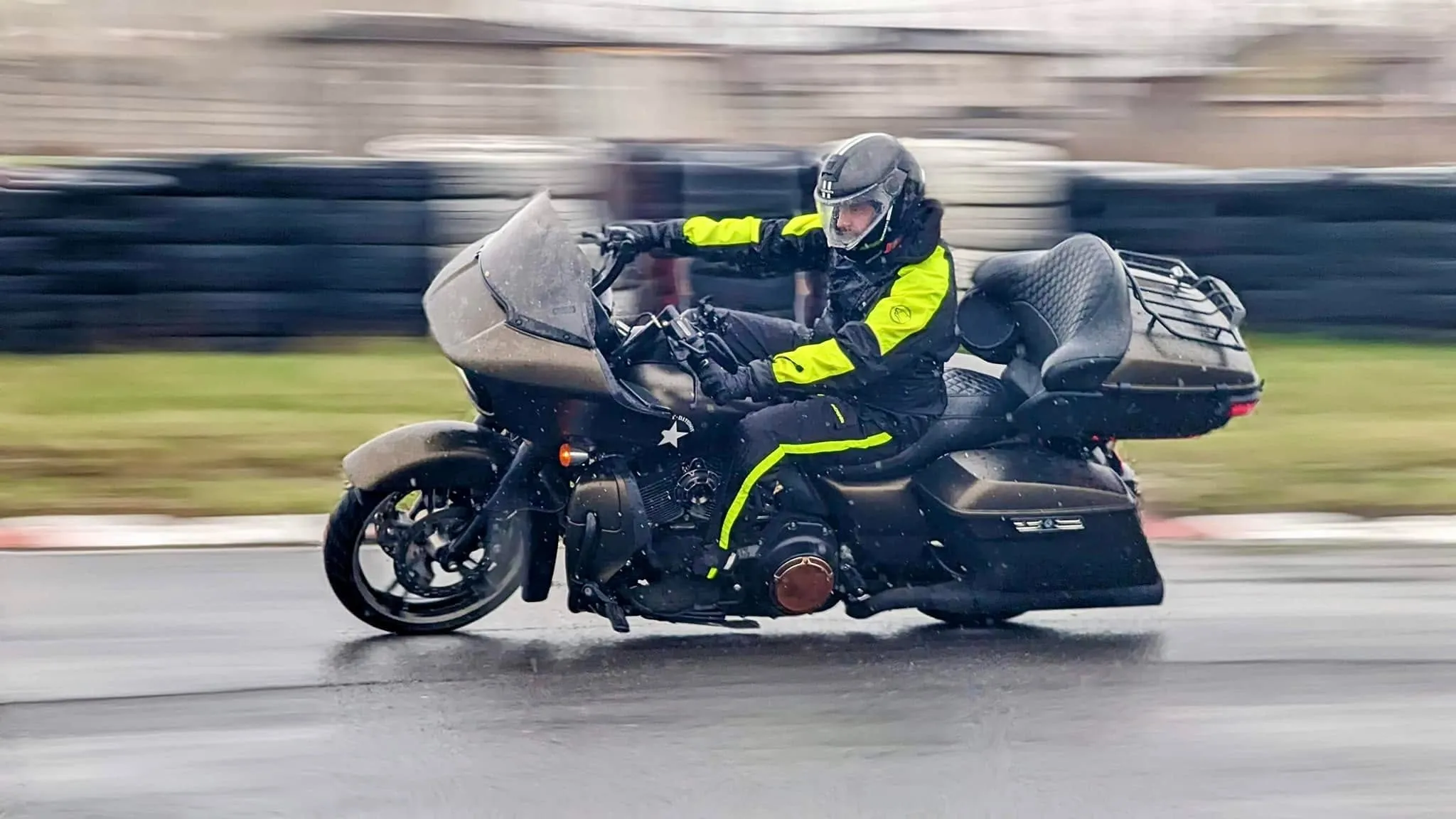 Un motociclist într-un costum negru cu galben neon conduce o motocicletă touring pe un circuit ud, în ploaie.