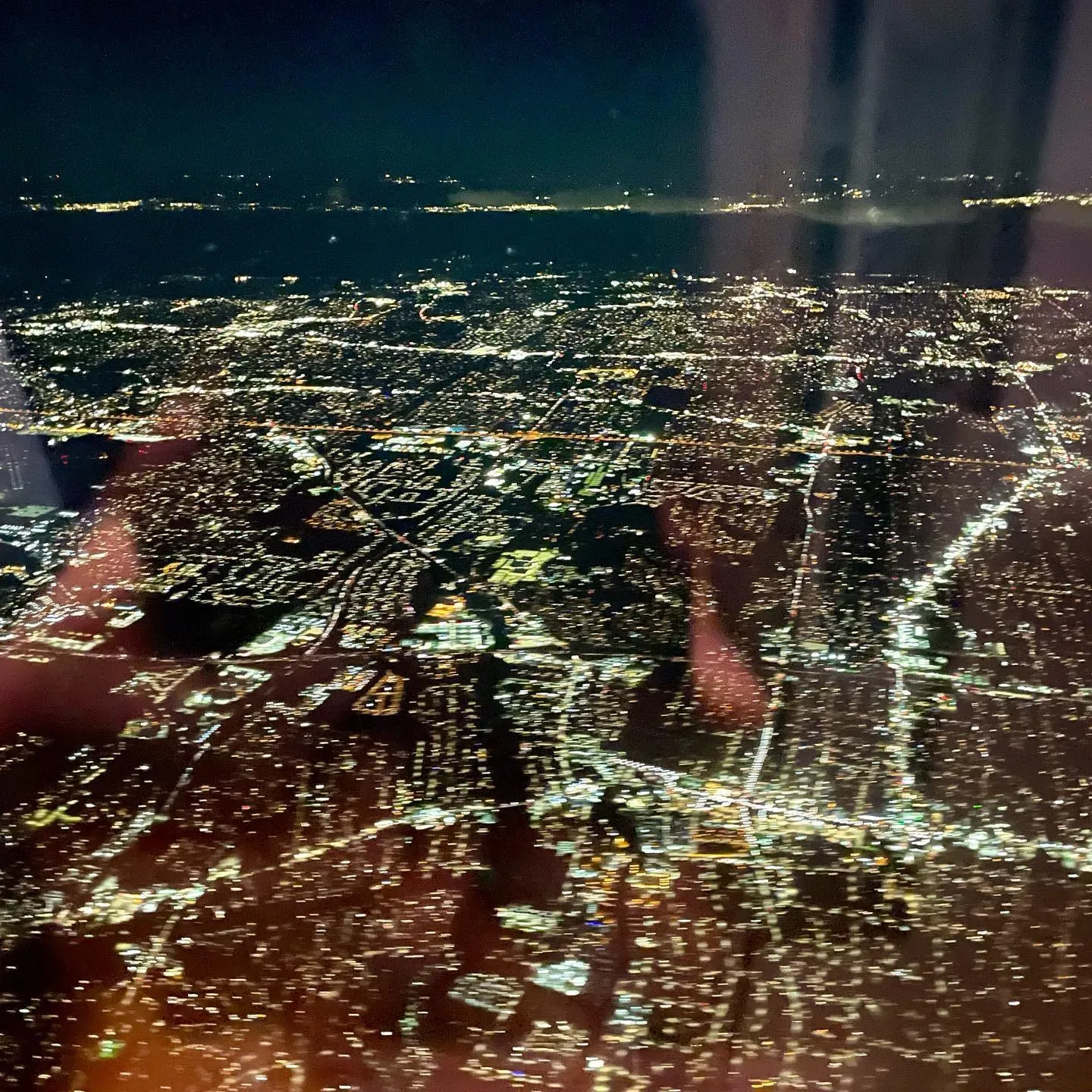 New York City glows at night beneath an airplane, with cabin reflections visible across the window.