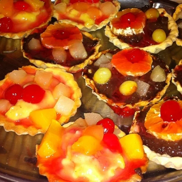 Colorful mini fruit tarts on a metal tray, topped with pineapple, cherries, oranges and grapes.