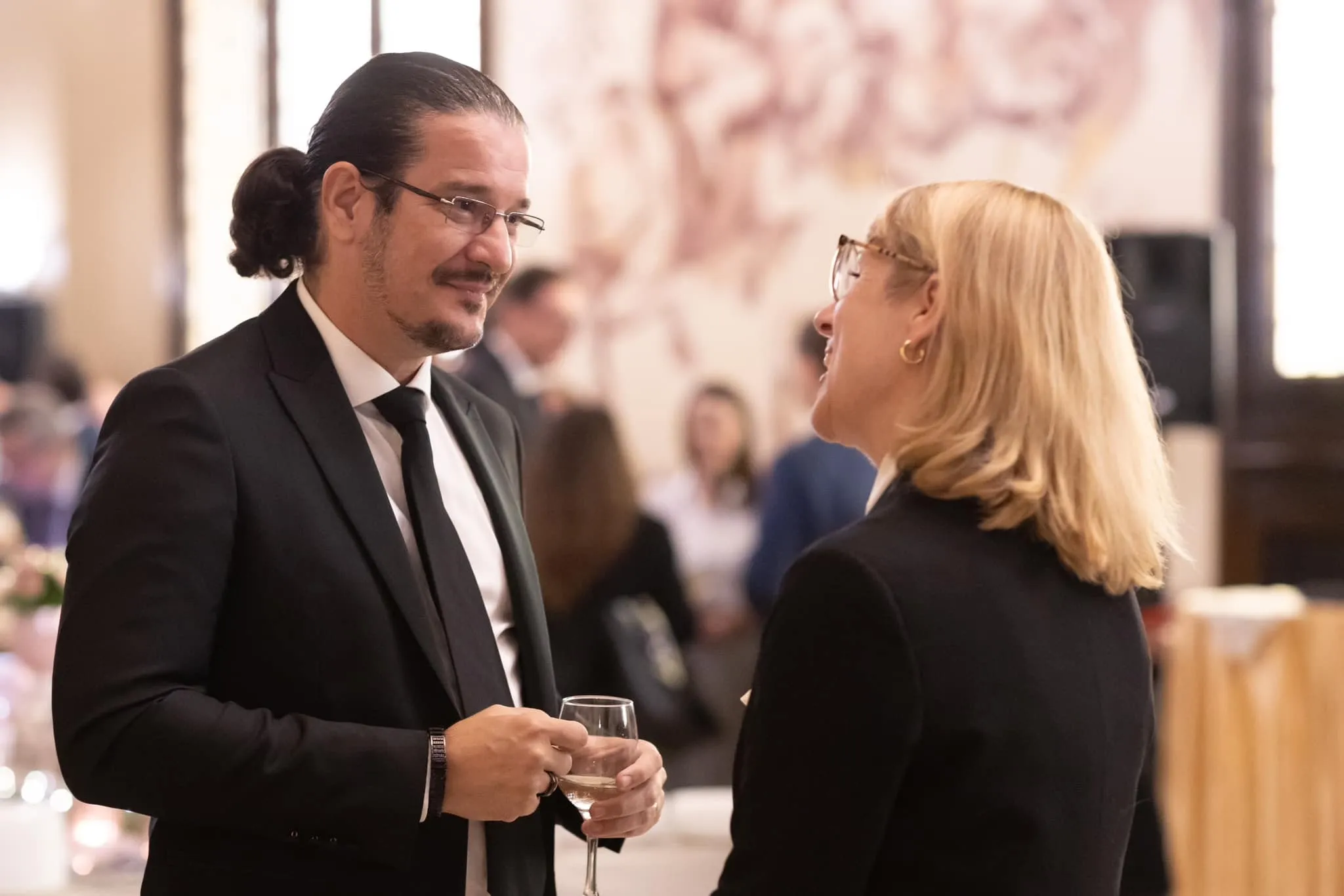 Un bărbat în costum negru și o femeie blondă îmbrăcată într-un sacou discută ținând în mână pahare cu vin, la o recepție oficială.