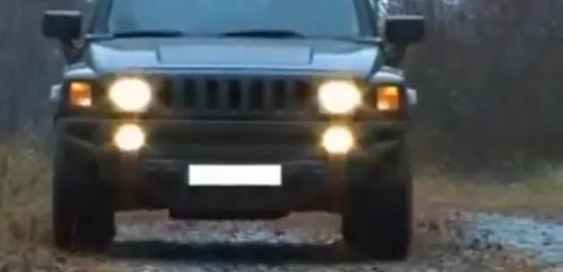 Un automobil Hummer cu farurile aprinse se apropie pe un drum neasfaltat.