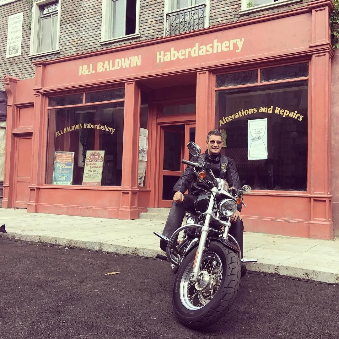 Un bărbat îmbrăcat într-o geacă neagră de piele stă pe o motocicletă în fața vitrinei roșii a magazinului J&J Baldwin Haberdashery