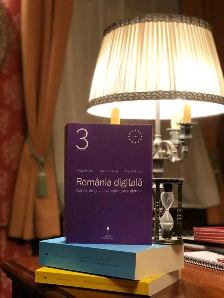 The third Digital Romania volume stands on two other books next to a lit table lamp and an hourglass.