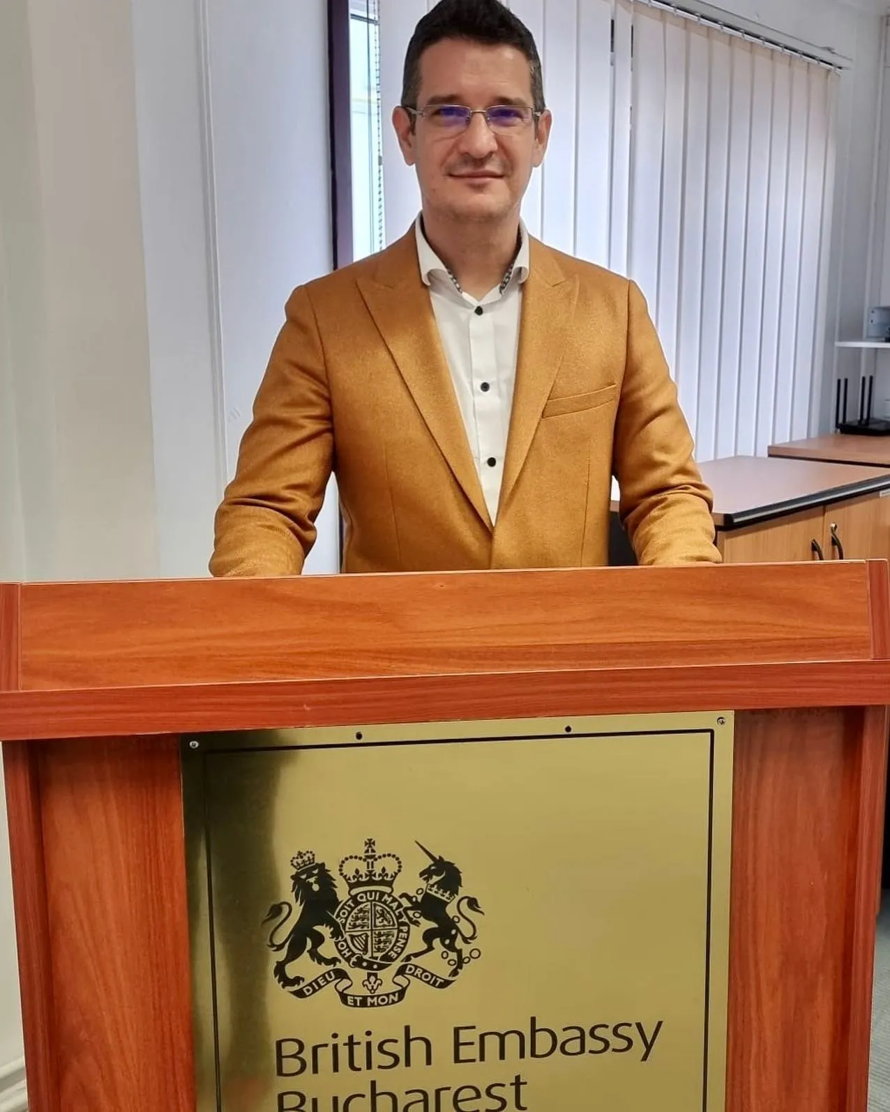 Sebastian Cochinescu se află în spatele unui pupitru pe care este afișată emblema Ambasadei Marii Britanii la București.
