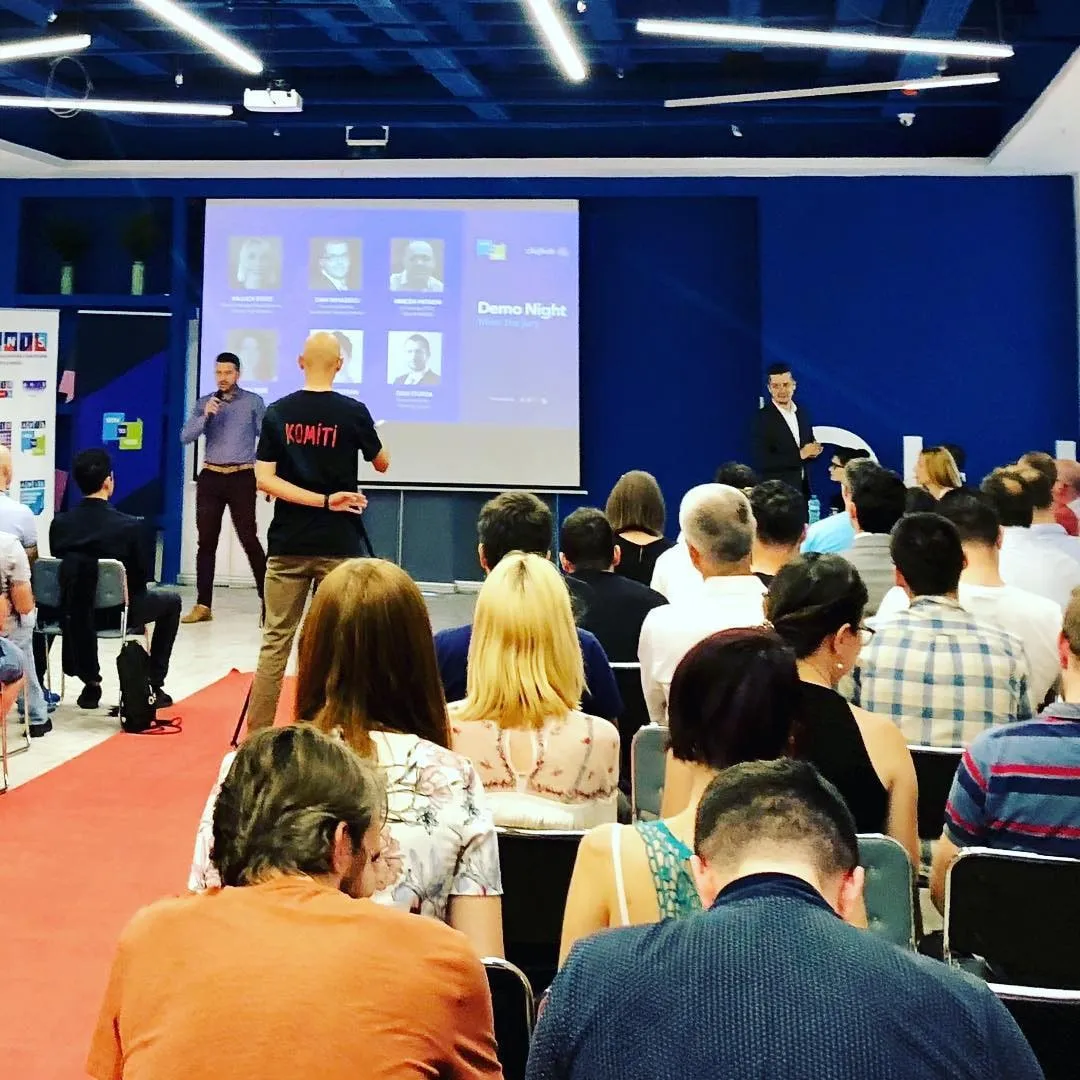 Publicul urmărește prezentatorii aflați pe scenă, sub un slide Demo Night, la un eveniment într-o sală cu pereți albaștri și mochetă roșie.