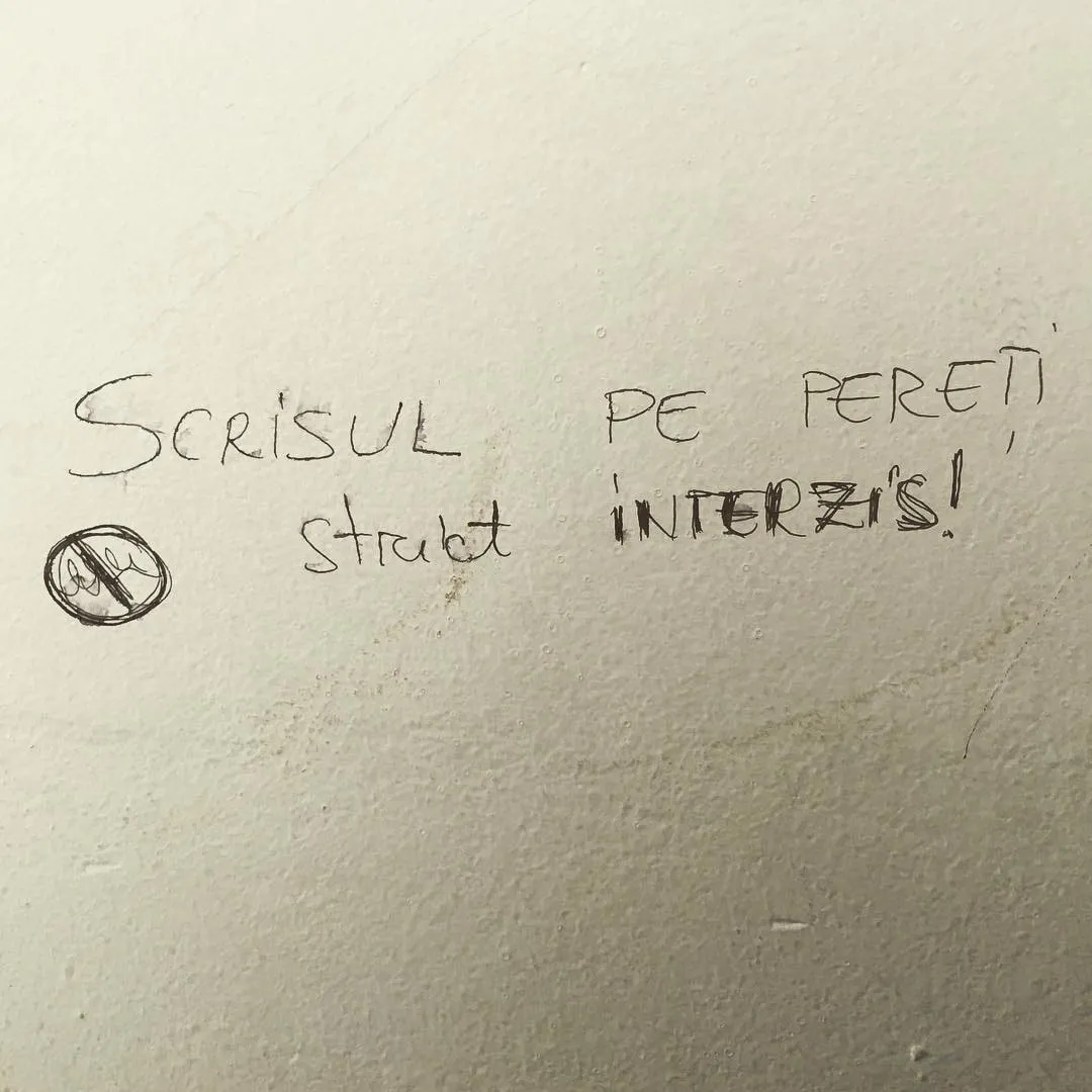 Handwritten Romanian graffiti on a plain wall reads Scrisul pe pereti, strict interzis beside a crossed-out pen symbol