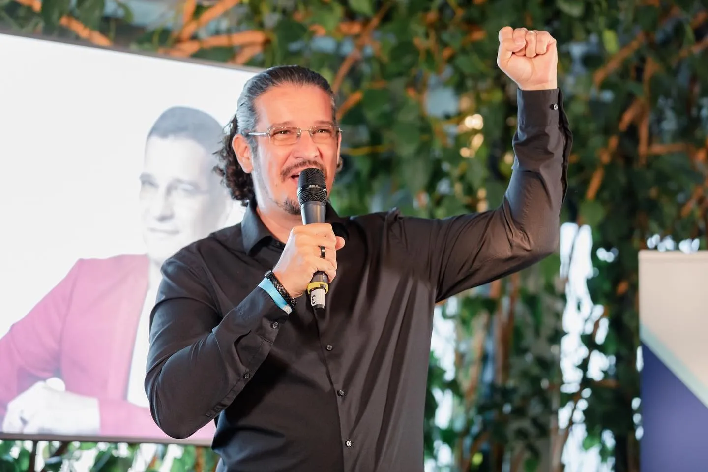 Man in a black shirt speaks into a microphone with his fist raised on stage, a portrait of himself projected behind him.