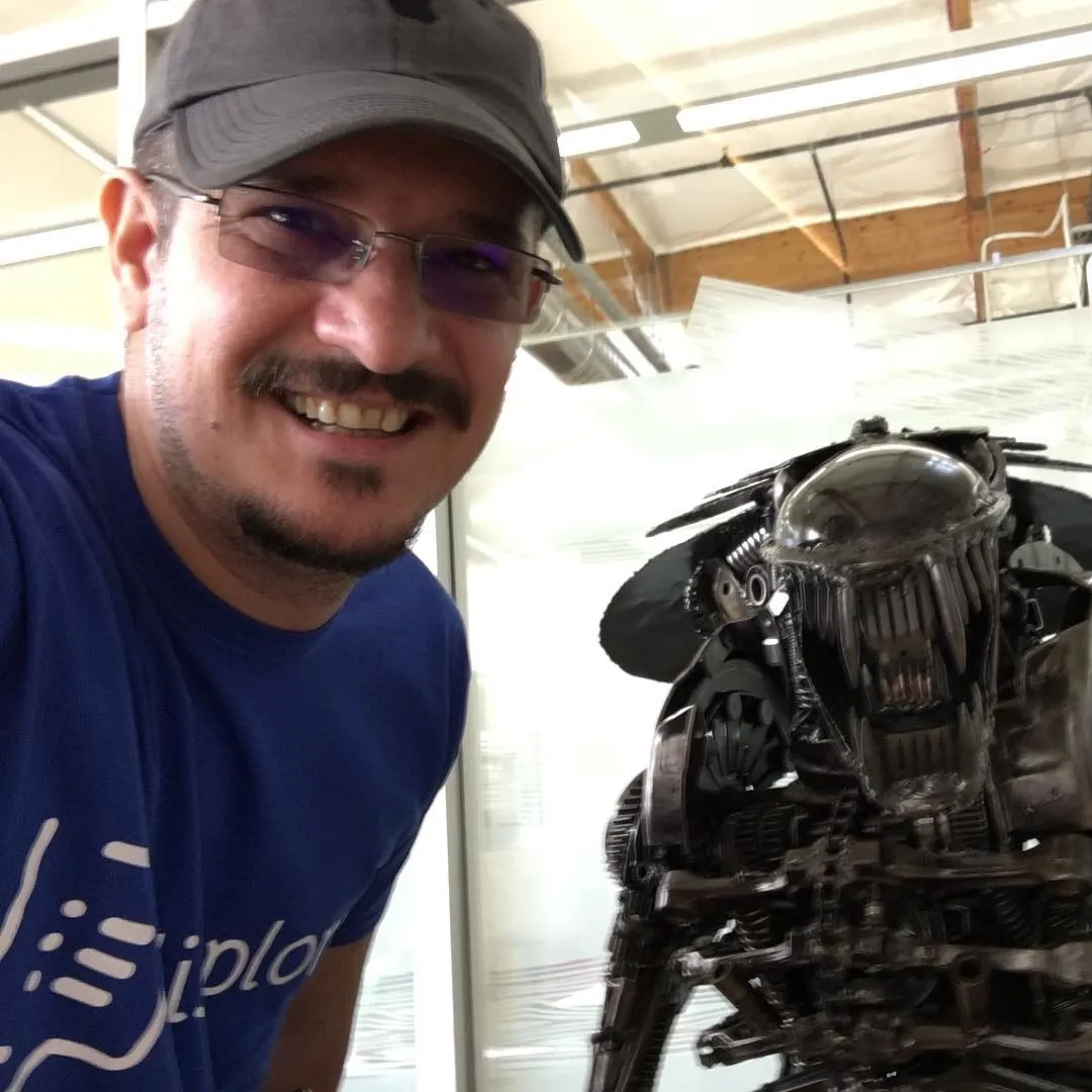 A smiling man in a blue shirt, glasses, and baseball cap poses beside a metal Predator sculpture