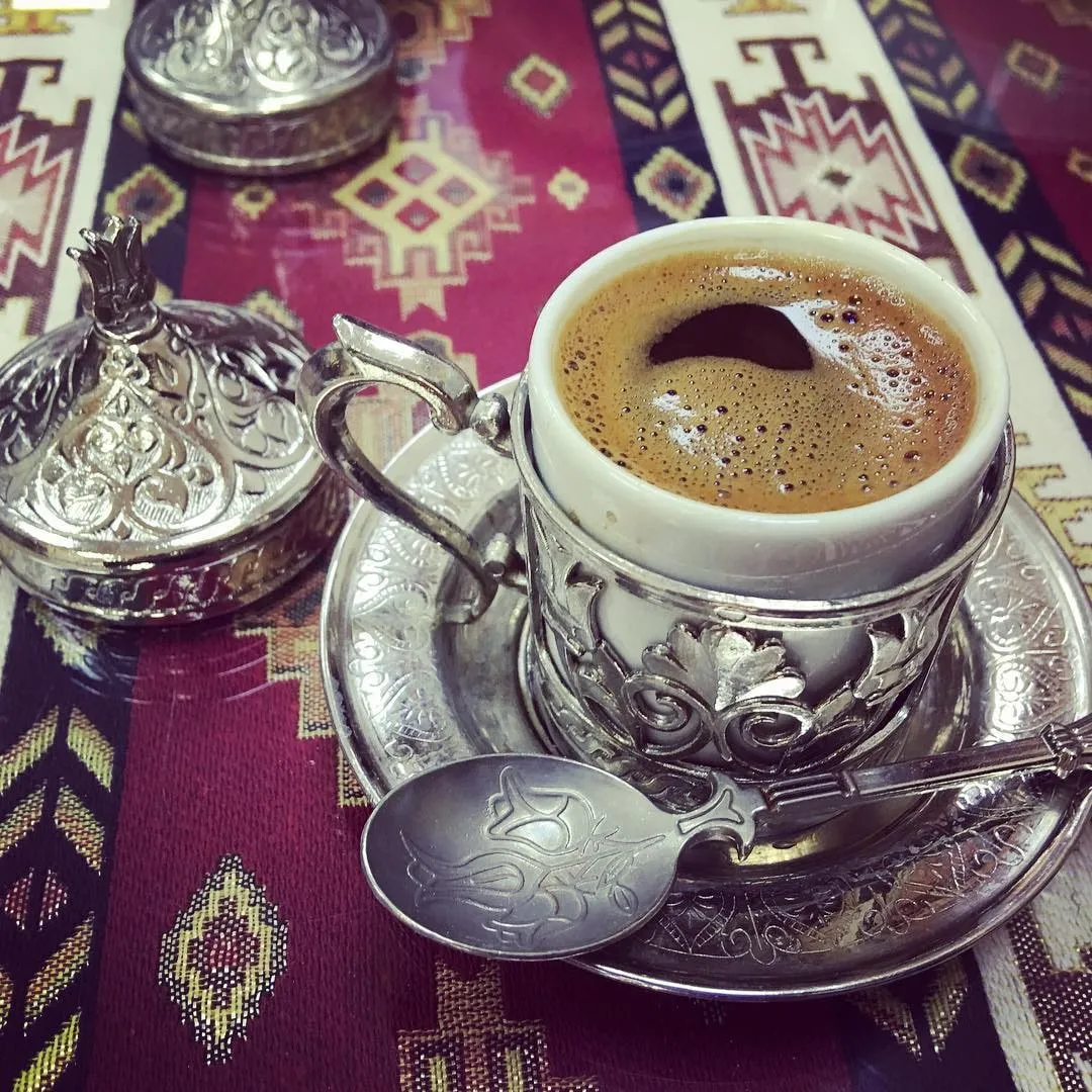 Cafea tradițională într-o ceașcă cu suport argintiu ornamentat și o linguriță, pe o față de masă cu model.