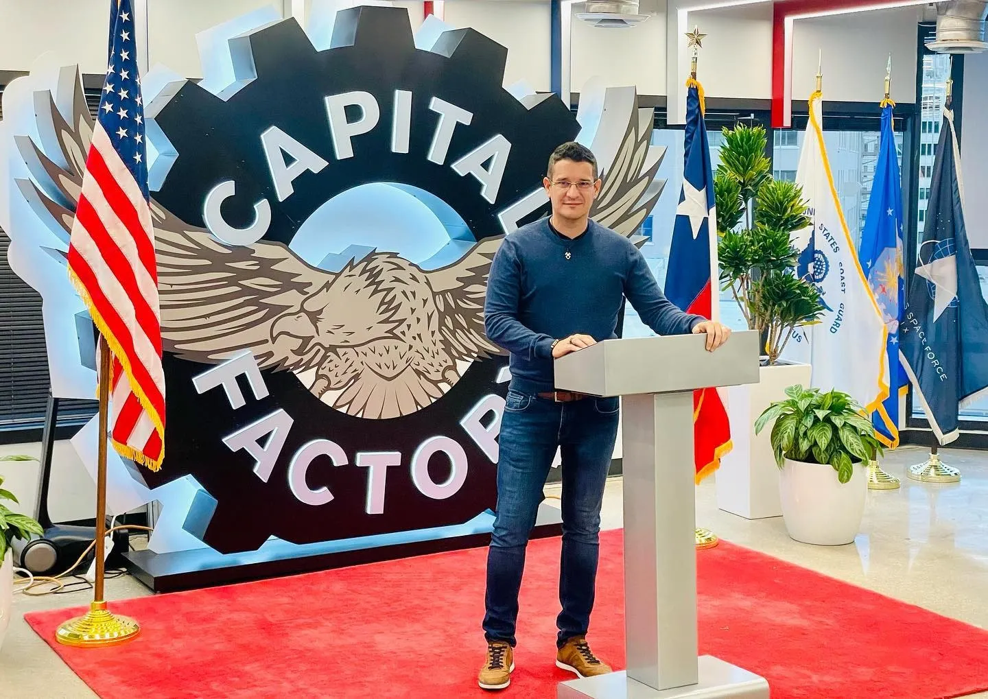 Sebastian Cochinescu stă la un pupitru, lângă emblema Capital Factory și mai multe steaguri.