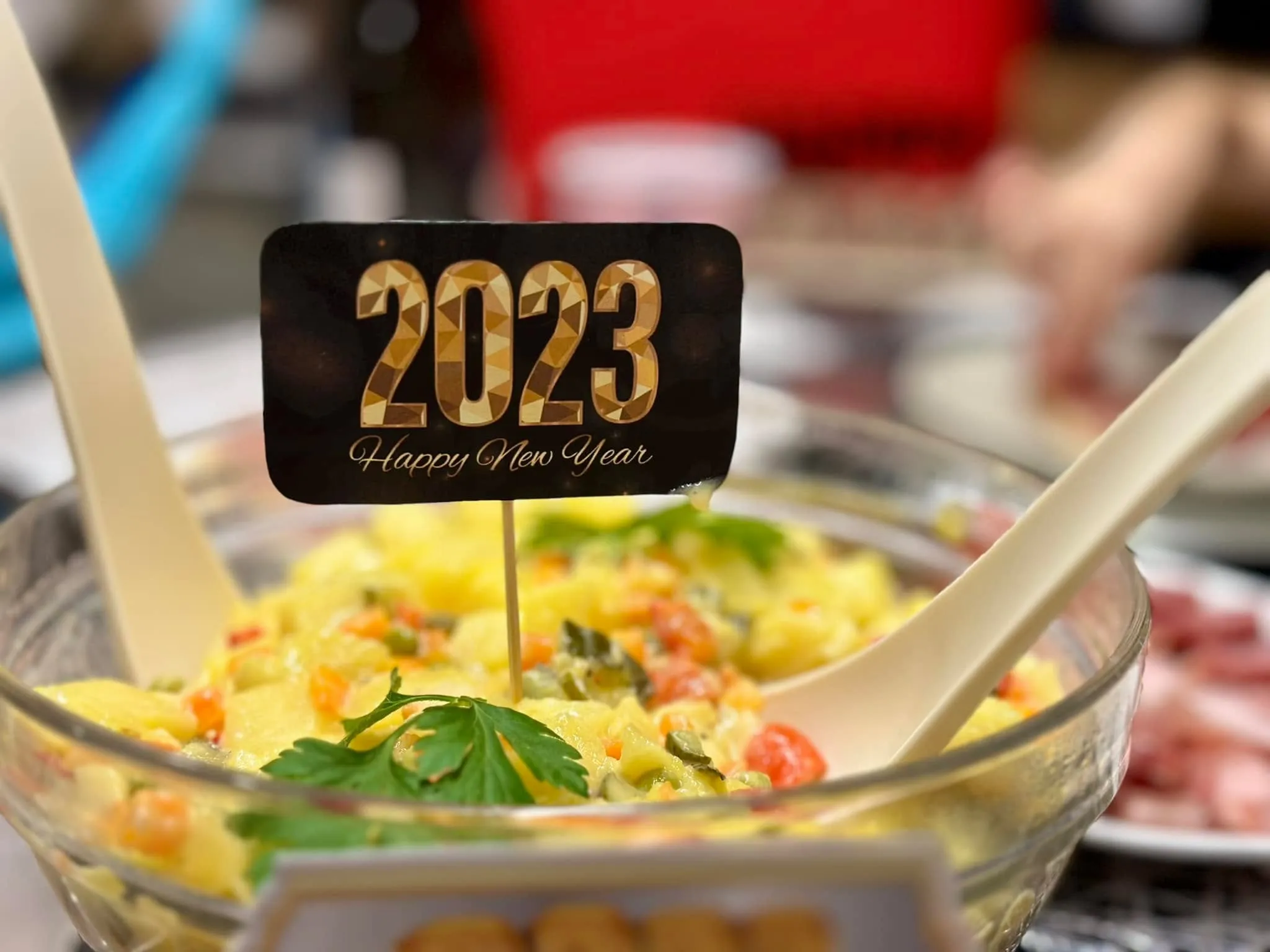 A black 2023 Happy New Year pick standing in a glass bowl of potato salad with serving spoons.