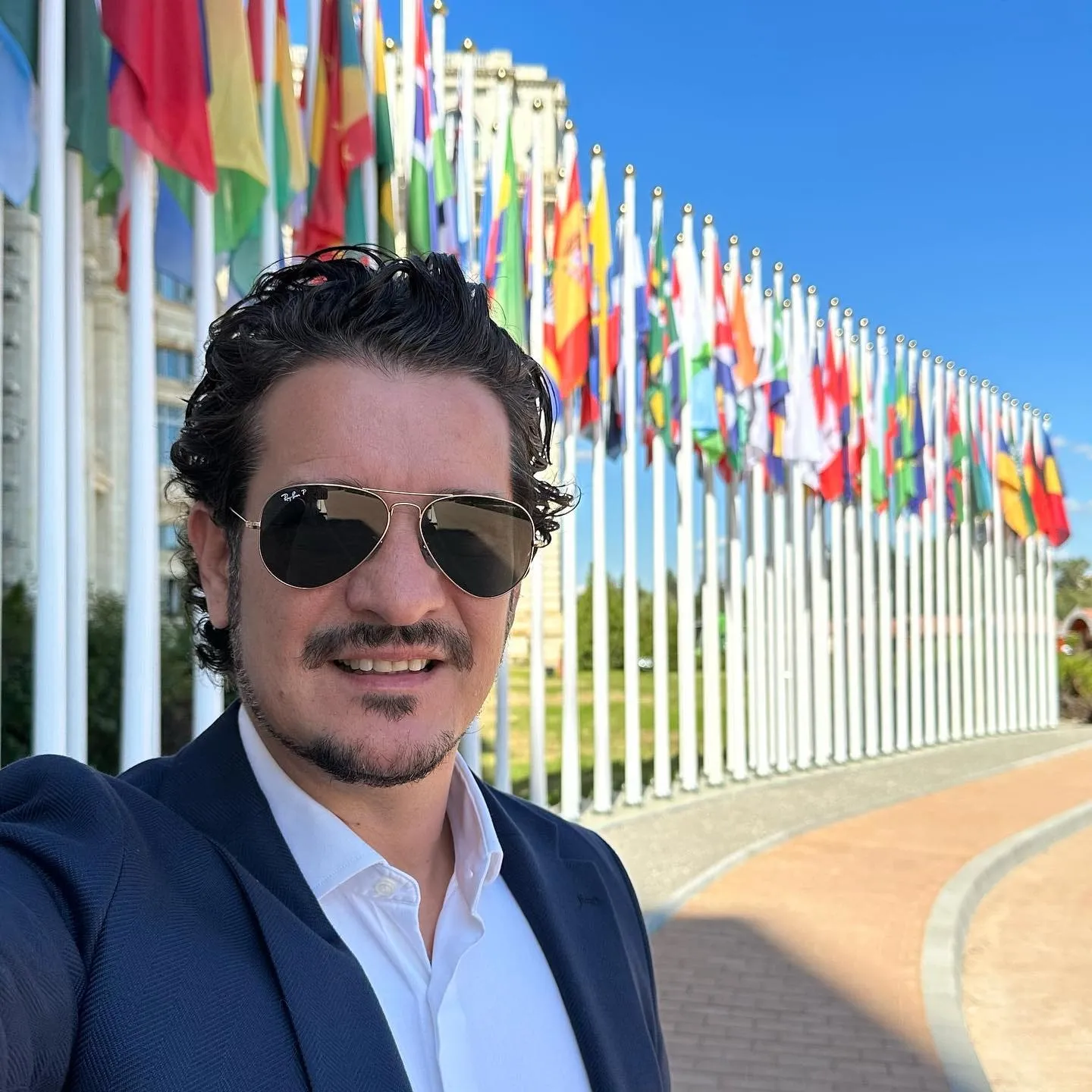 Un bărbat cu ochelari de soare și sacou albastru își face un selfie în fața unor șiruri de steaguri internaționale.