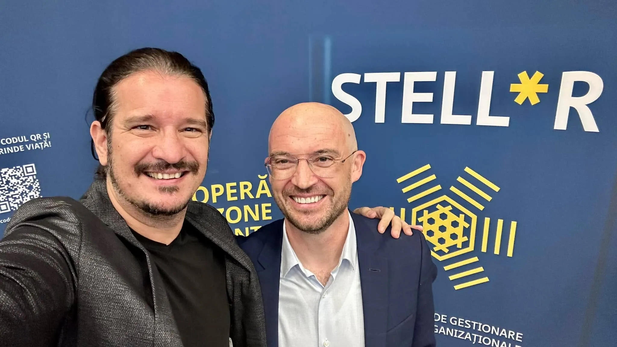 Doi bărbați zâmbitori își fac un selfie în fața unui panou albastru STELLR, cu un logo geometric auriu.