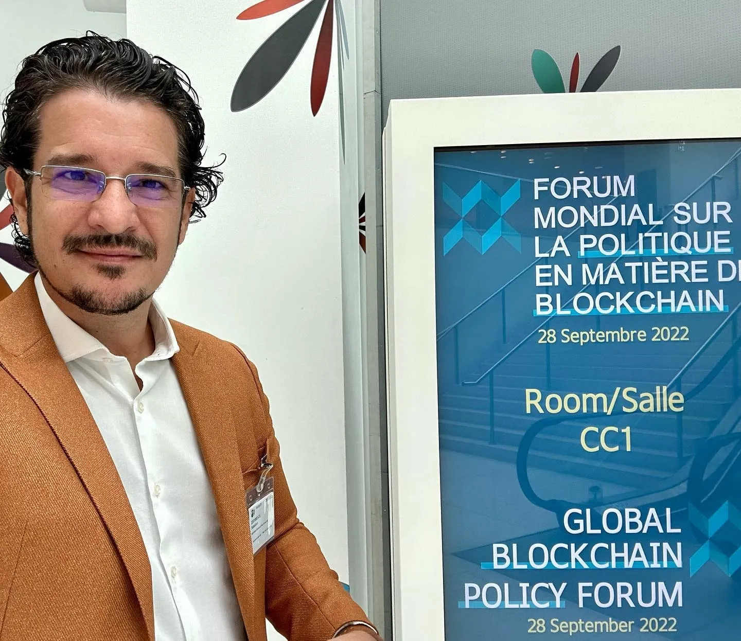 Un bărbat într-un sacou maro stă lângă un panou Global Blockchain Policy Forum 2022.
