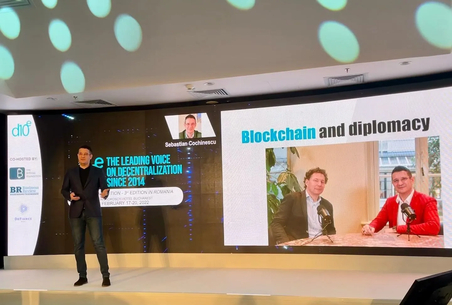 Sebastian Cochinescu vorbește pe scenă, lângă un slide de prezentare intitulat Blockchain și diplomație.