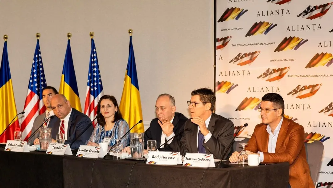 Paneliști, inclusiv Sebastian Cochinescu, stau la o masă de conferință cu microfoane, sub steagurile României și SUA, la un eveniment Alianța.