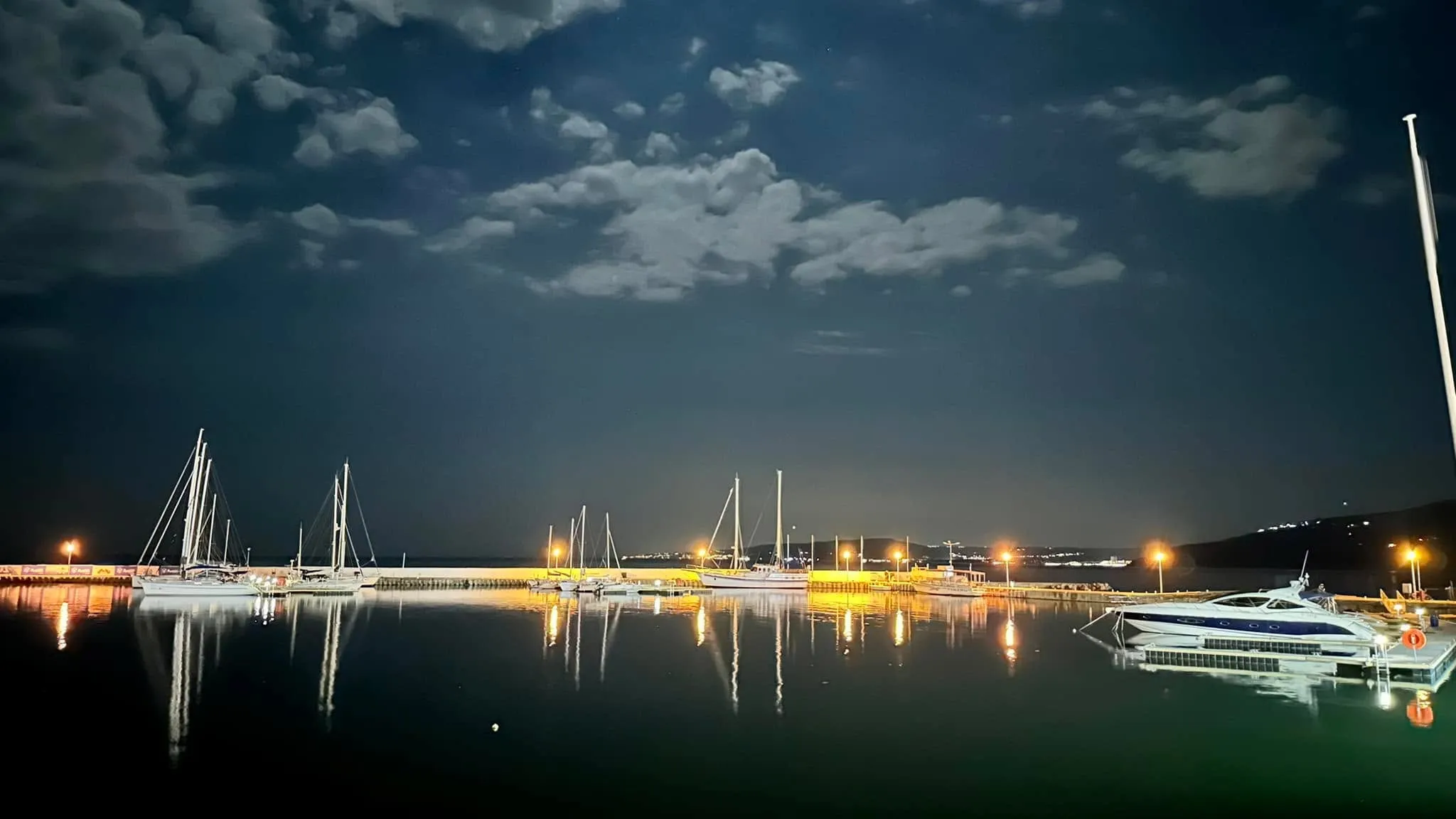 Sailboats and a yacht docked at a marina at night, with warm lights reflecting on calm water under a cloudy sky.