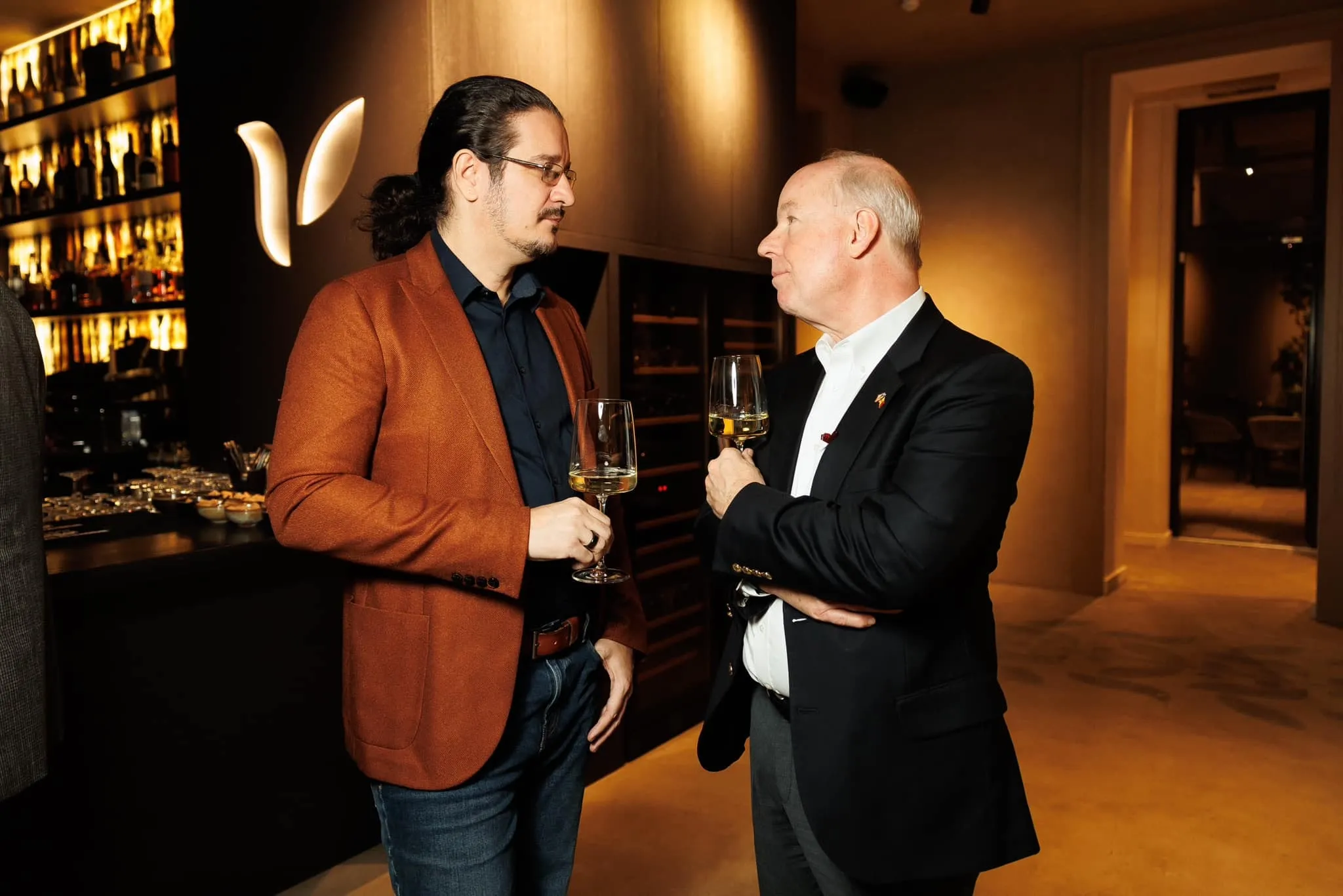 Sebastian Cochinescu și ambasadorul SUA Kent D. Logsdon discută, ținând în mână pahare cu vin, într-un lounge.