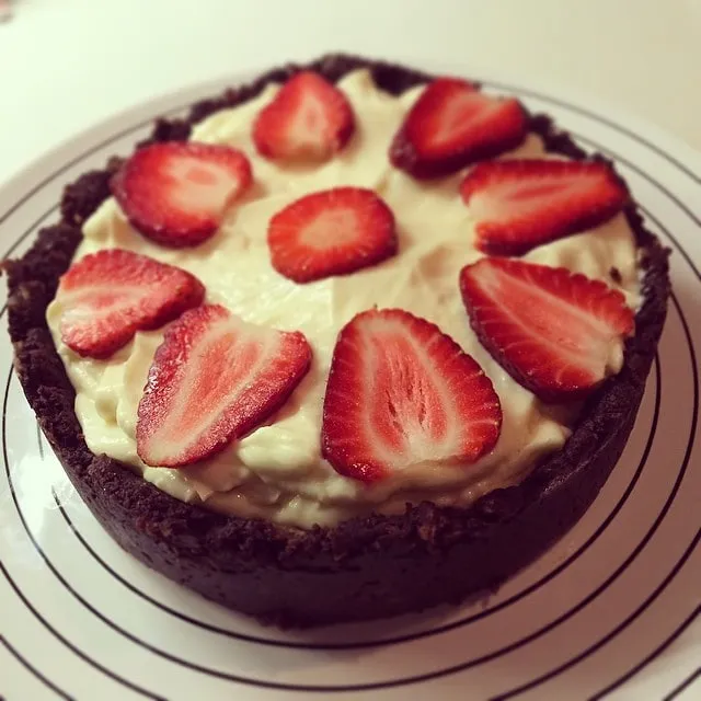 Round chocolate-crusted cake with white frosting and strawberry slices on a plate with black stripes.