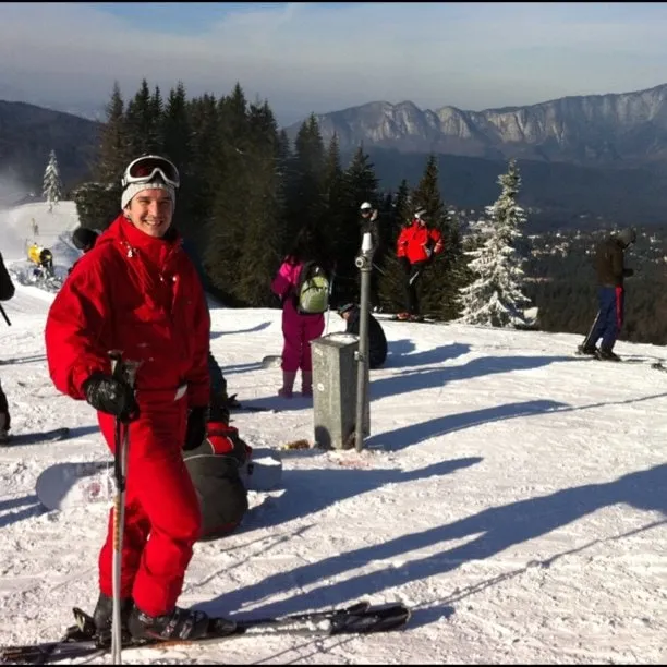 Bărbat în combinezon roșu de schi pe o pârtie la Clăbucet, cu pini, alți schiori și munți în fundal.