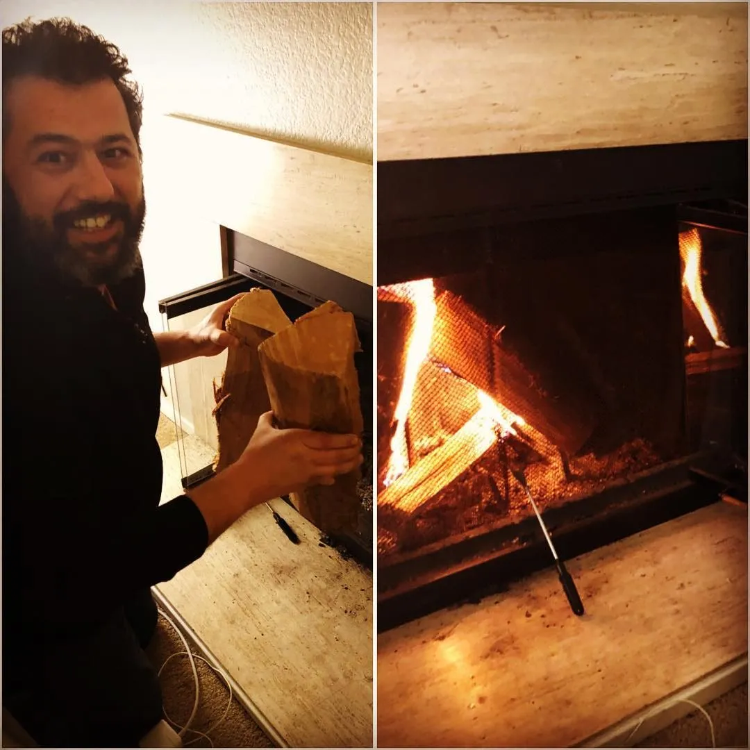 Alăturare cu un bărbat bărbos și zâmbitor ținând lemne de foc, lângă un șemineu aprins, cu un vătrai pe vatră