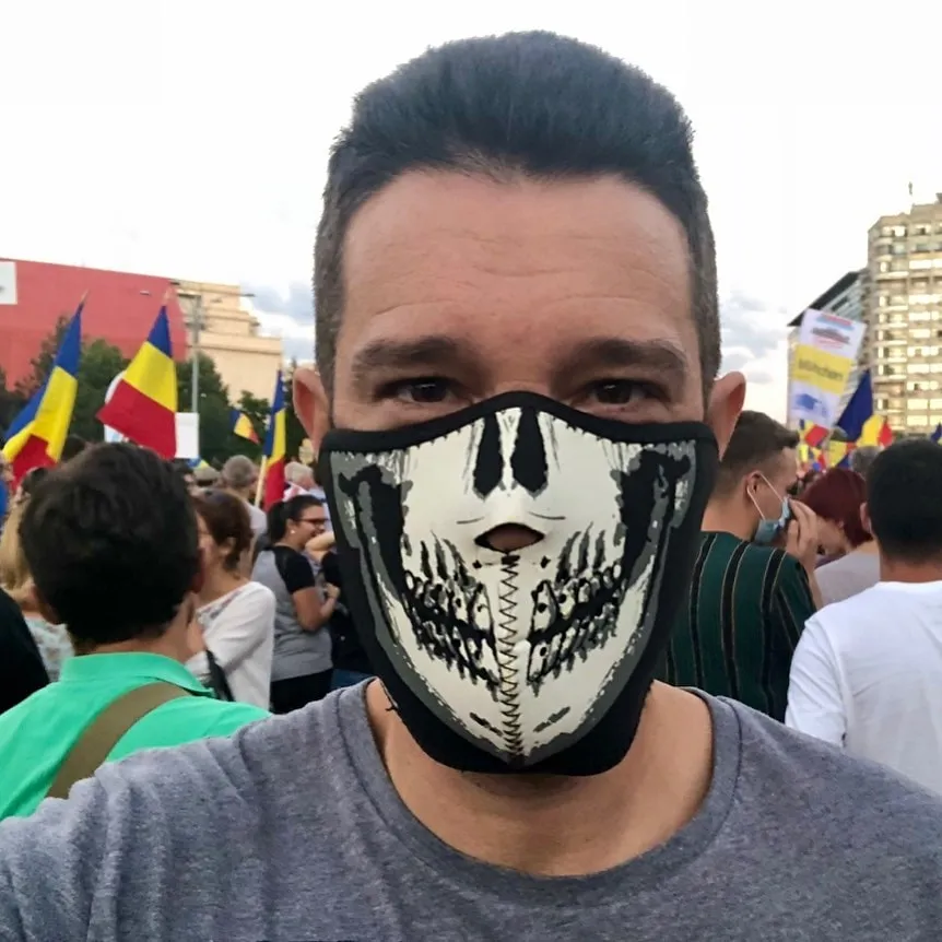 Sebastian Cochinescu poartă o mască cu cranii la un protest aglomerat, printre steaguri ale României.
