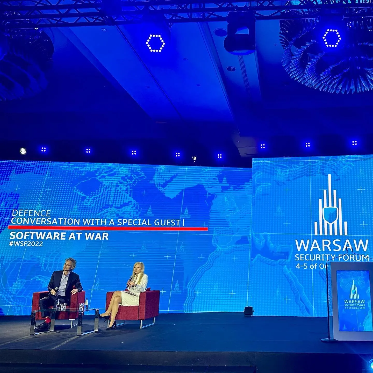 Alex Karp și un moderator stau pe scenă la Warsaw Security Forum, sub un ecran pe care scrie Software at War.