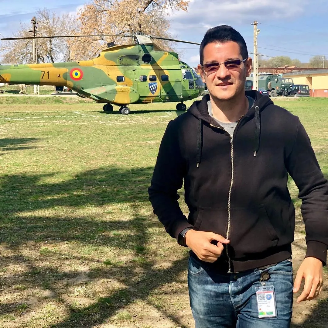 Un bărbat cu ochelari de soare și un hanorac negru stă pe iarbă în fața unui elicopter militar românesc camuflat.