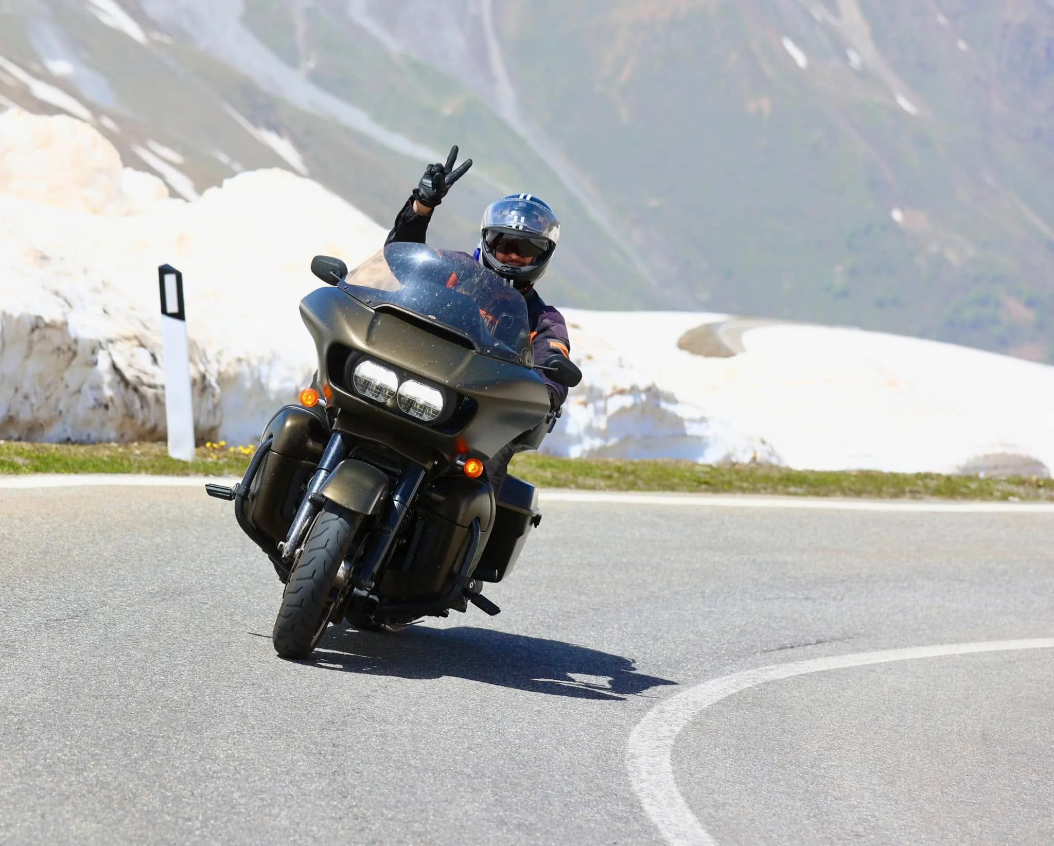 A motorcyclist in a helmet rides a black touring motorcycle on a mountain road lined with snow banks.