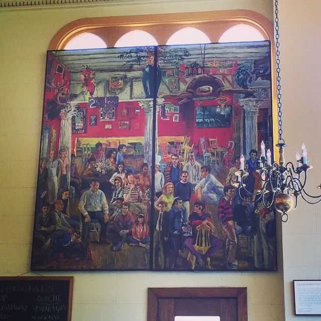Pictură murală mare cu o scenă de cafenea plină de oameni, pe un perete cu ferestre arcuite.