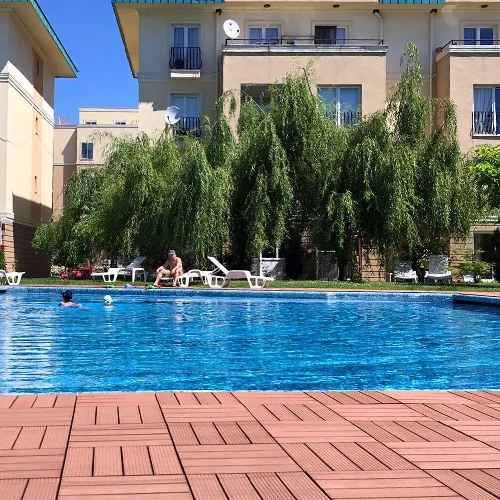 Piscină în aer liber cu oameni care înoată, un bărbat pe un șezlong și blocuri bej în spatele unei sălcii